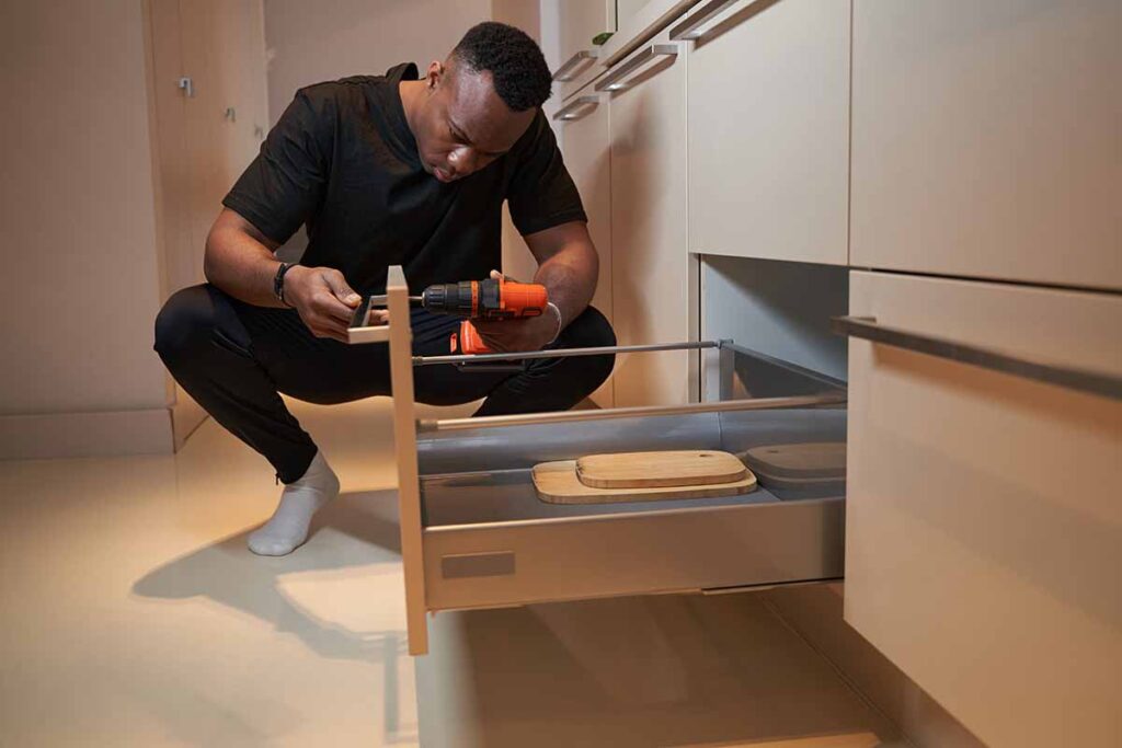 Concentrated mixed-race man sitting on kitchen near opened drawer and fixing its handles with screw gun