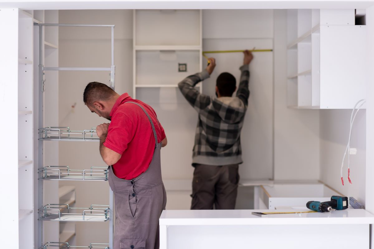 Hire Professional Cabinet Installer Services | Dr. Cabinet 2 Two workers are installing white kitchen cabinets; one measures the wall, the other adjusts pull-out shelves.
